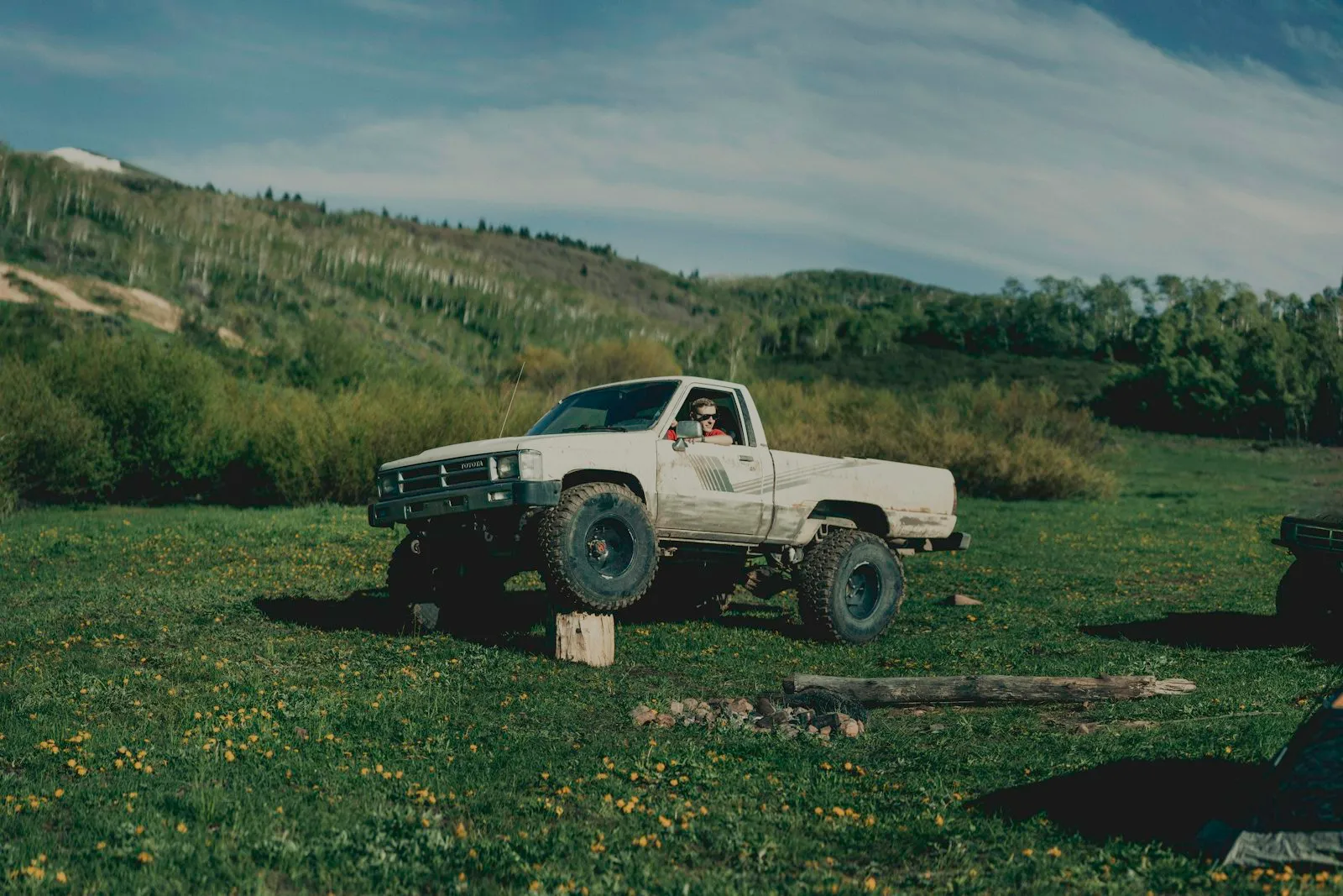 Photo of a vintage Toyota truck in a heroic pose on a log. Situated in the middle of a mountain meadow with a tent off to the side.